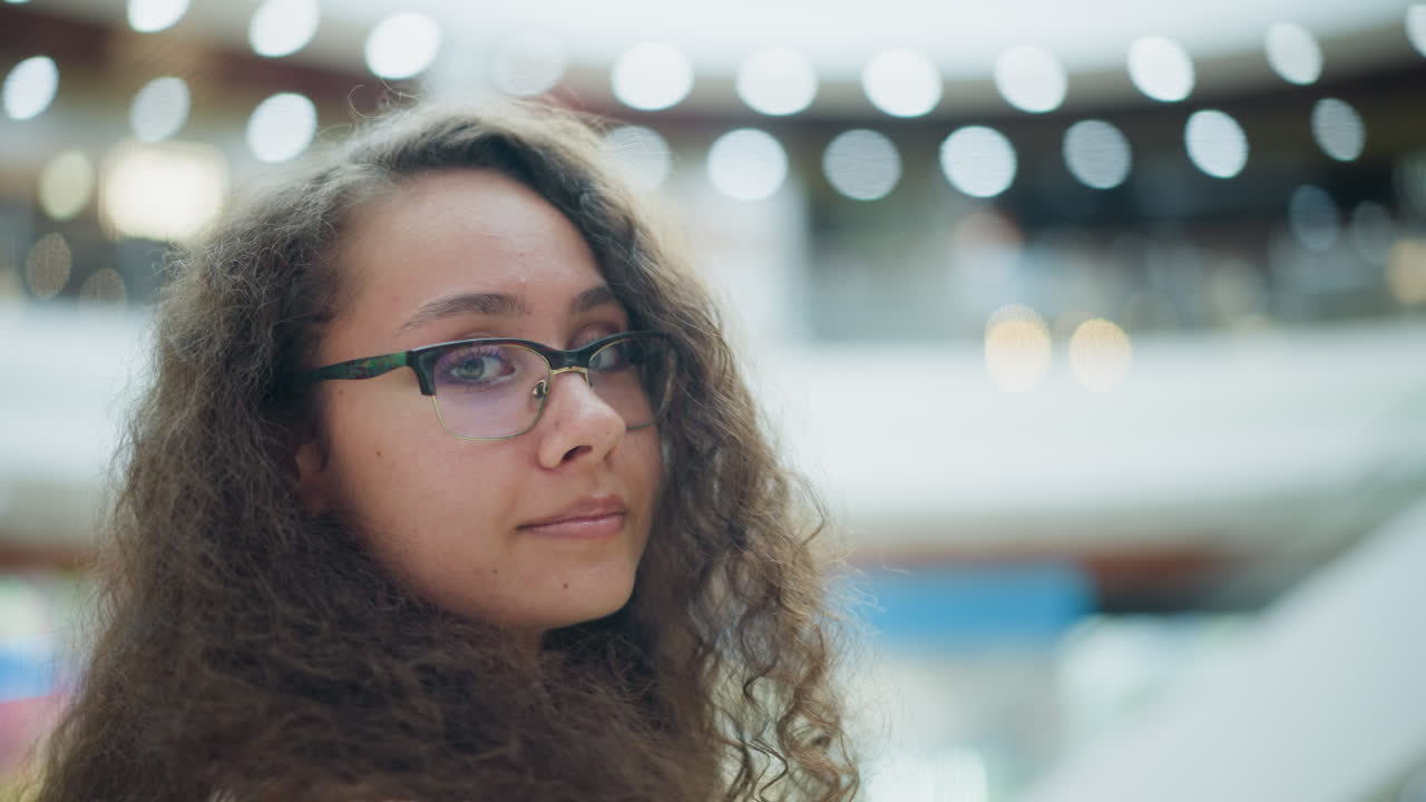 Young woman with curly hair and glasses standing in a well-lit mall, looking towards the camera with a soft, gentle smile, the background features beautiful bokeh lighting