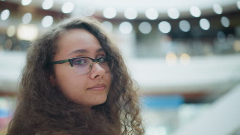 mujer joven con cabello rizado y gafas de pie en un centro comercial bien iluminado, mirando hacia la cámara con una sonrisa suave y suave, el fondo cuenta con una hermosa iluminación bokeh