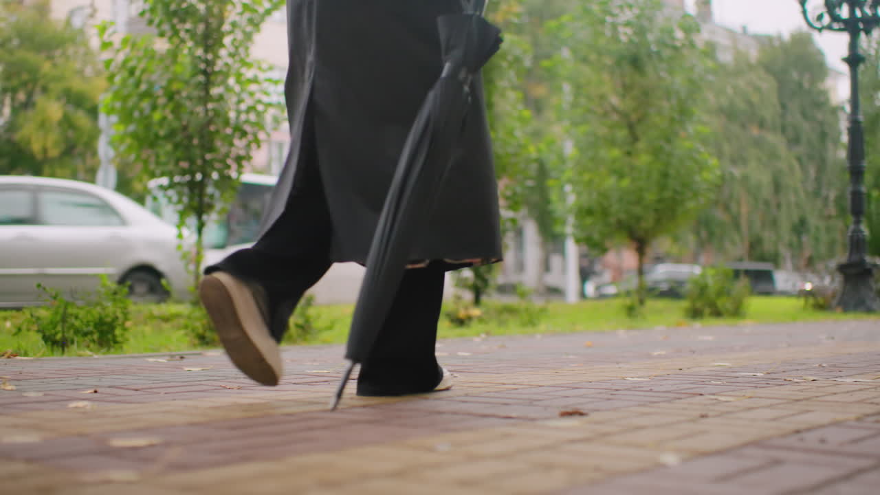Woman in long black coat holding closed umbrella walks on wet pavement during overcast autumn day, close-up of legs and coat hem, emphasizing urban atmosphere, solitude, moody lifestyle