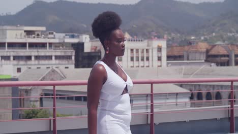 in a white dress, a young black girl stands on a rooftop with a view of the city