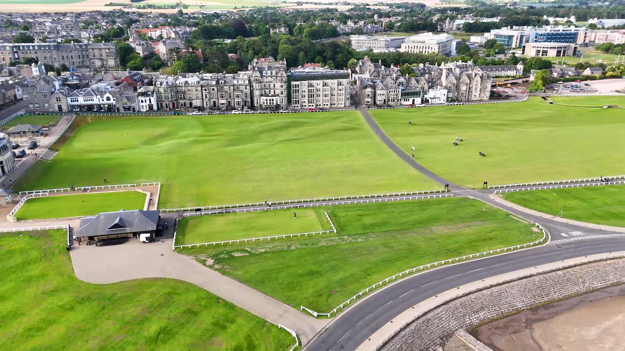 Drone camera smoothly pans above lush green golf course, revealing Victorian brick architecture and surrounding town under bright daylight in St Andrews, Scotland