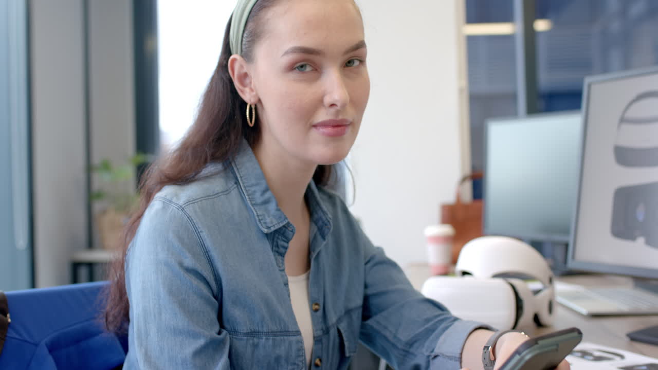 Using smartphone, woman in denim shirt working at office desk with VR headset