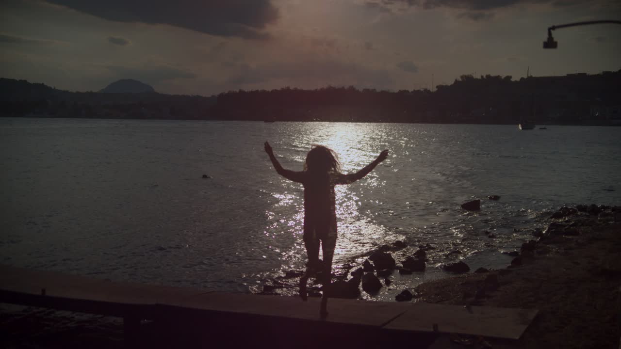 Silueta de mujer bailando sirtaki en un muelle frente al mar al atardecer.
