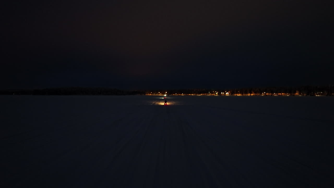 Drone following of a snowmobile onr a lake, toward a town,, winter night in Lapland