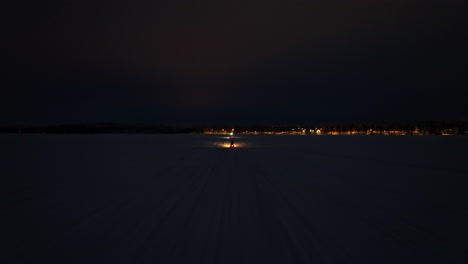 drone following of a snowmobile onr a lake, toward a town,, winter night in lapland