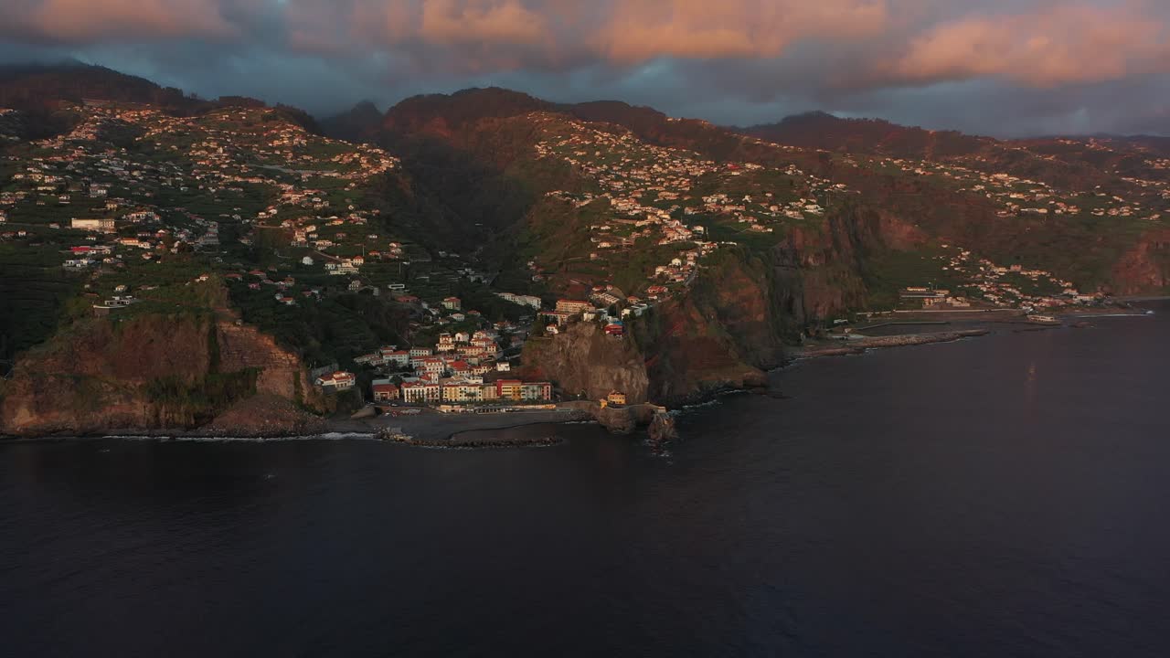 Slow drone shot moving backwards. The coastline of Ponta do Sol in Madeira during a dramatic sunset with a storm coming in over the peak of the mountain.