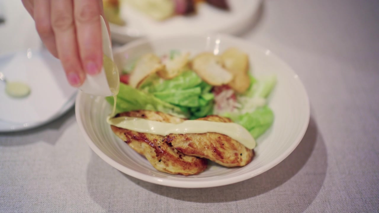 Nærbilde av en kvinne som klemmer kremet dressing fra en liten hvit sausmugge over grillet kylling servert i en hvit bolle med Caesar-salatingredienser, på en lys duk