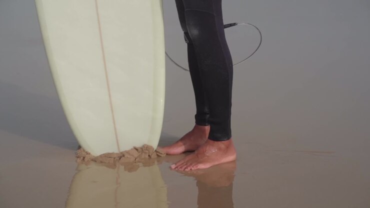 Inquadratura dal basso di un surfista in muta, a piedi nudi sulla sabbia bagnata della spiaggia con la tavola da surf bianca, ripresa a mano