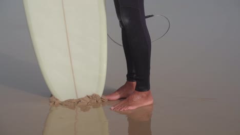 ángulo bajo de surfista en traje de neopreno, descalzo en el suelo de la playa de arena húmeda con tabla de surf blanca, tiro de mano