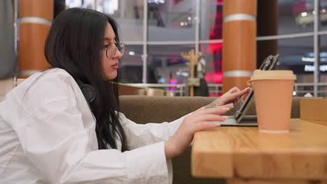 side view of young woman with long hair wearing glasses tapping wooden table thoughtfully while operating tablet beside coffee cup in cozy lounge with ambient lights and blurred modern background