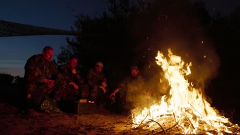 a large bonfire flame, behind it military men in camouflage uniforms are sitting, talking and warming themselves at night by the hearth