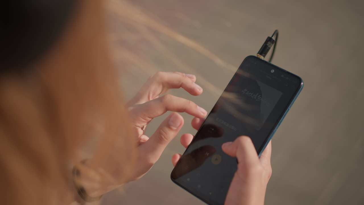 Primo piano di una donna che tocca lo smartphone con le cuffie sul marciapiede in città, controllando la playlist tra un passo di danza e l'altro; mani e schermo visibili, cavo degli auricolari e braccialetto in vista, capelli lunghi mossi dalla brezza, atmosfera calda