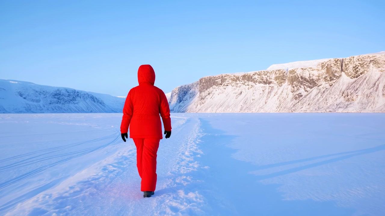 Rear view shows a lone figure in vivid red outerwear moving steadily through deep snow, with existing tracks leading forward, snowy ridges ahead, and crisp daylight casting long shadows.