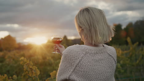 Eine-Frau-Mit-Einem-Glas-Rotwein-In-Der-Hand-Steht-Vor-Dem-Hintergrund-Eines-Weinbergs,-In-Dem-Die-Sonne-Wunderschön-Untergeht