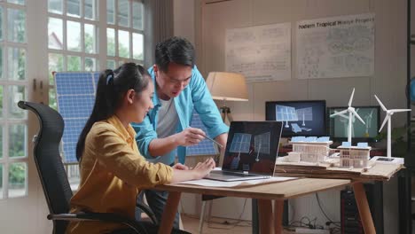 asian man and woman discuss about solar cell on laptop next to the model of a small house with solar panel and wind turbine