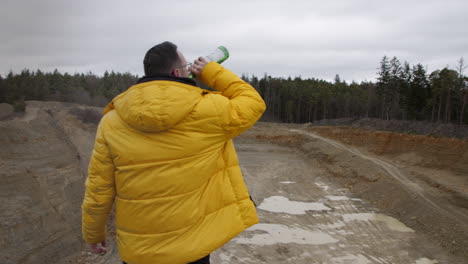 joven con abrigo amarillo bebiendo de una botella de whisky en la naturaleza rural