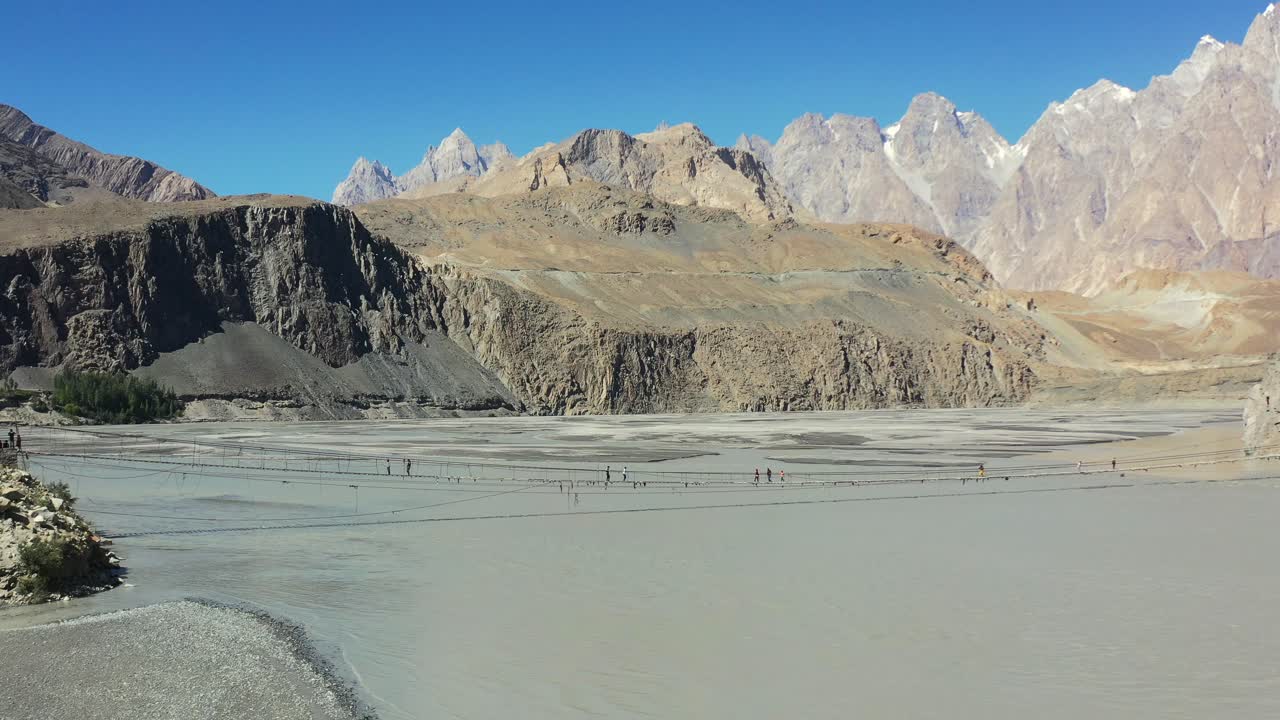 dron aéreo panorámico a la derecha que revela el famoso puente hussaini en hunza pakistán y passu cones montañas en la distancia mientras los turistas cruzan con cautela el peligroso puente sobre el río gris de ritmo rápido