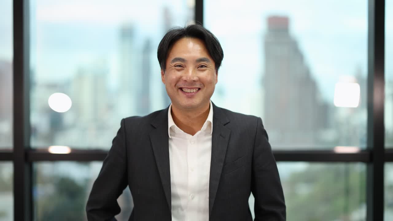 Asian businessman in suit smiles confidently, natural daylight, cityscape background, steady camera, positive mood