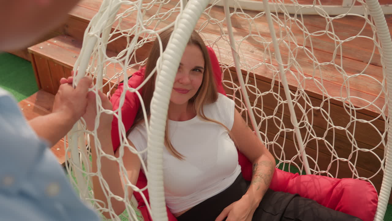 Donna che si rilassa su un'altalena sospesa, cuscino rosso, terrazza sul tetto, pavimentazione in legno, mano di un accompagnatore che sistema il telaio in vimini, calda luce del sole, top bianco casual, gonna scura, sorriso spontaneo, sguardo sereno, appuntamento intimo