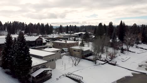 Snowy-American-small-town-in-winter-with-slow-moving-cars,-houses,-shops-and-surrounding-trees-in-McCall,-Idaho,-USA---Aerial-drone-rising