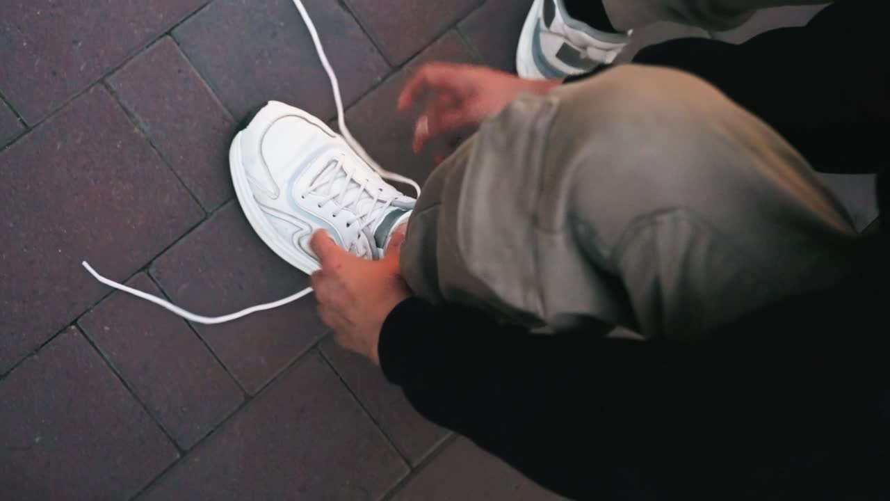 Primo piano dall'alto di mani che allacciano lacci bianchi di scarpe da ginnastica, mentre regolano il nodo sul marciapiede. Abbigliamento casual e postura concentrata, una routine spontanea di strada prima del pendolarismo, sotto una morbida luce mattutina. Si notano la suola strutturata e lunga.