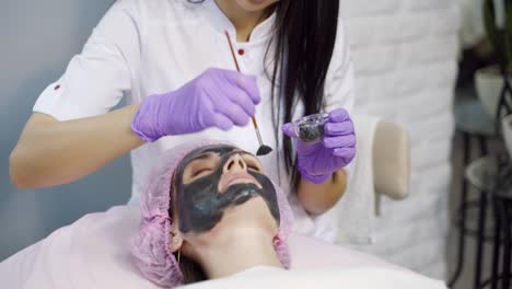 an experienced cosmetologist carefully applies a black cleansing mask to a woman's face in a beauty salon. close-up. industry of beauty.