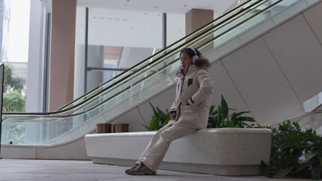 asian female puts on white headphones and turns on music sitting on a bench at the mall