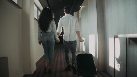 Sweet-couple-pulling-suitcase-in-corridor.-Young-couple-holding-hands-in-hotel