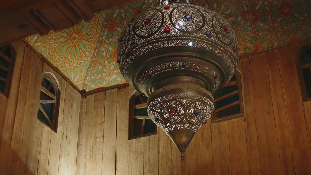 lanterna in ottone traforato con pietre preziose appesa a un soffitto con motivi zellige dipinti, con pareti in legno e finestre ad arco in un riad marocchino