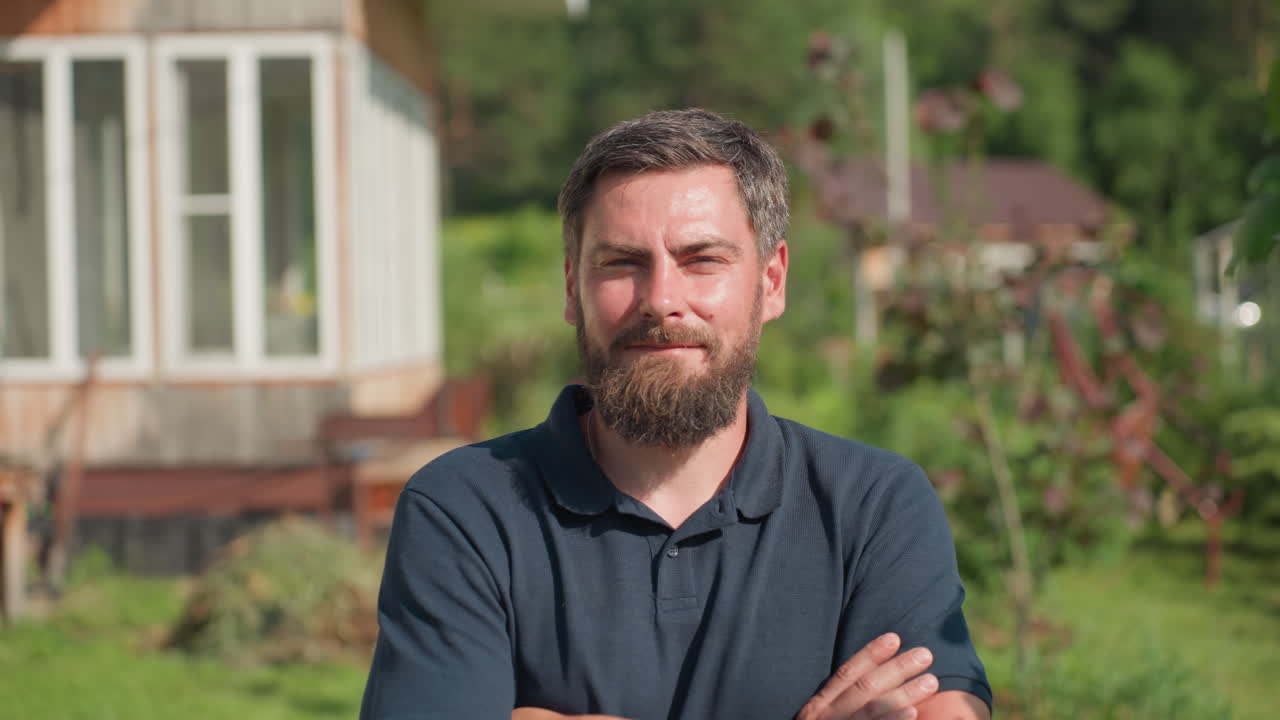 man turning and folding arm while looking at camera, leaves swaying beside him, shadow cast on shirt, rural house background, sunlight creating calm reflective mood on farm property