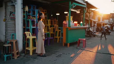 young woman is standing near a vibrant juice stand filled with colorful plastic crates of fruit, bathed in the warm glow of the setting sun in a bustling tropical city