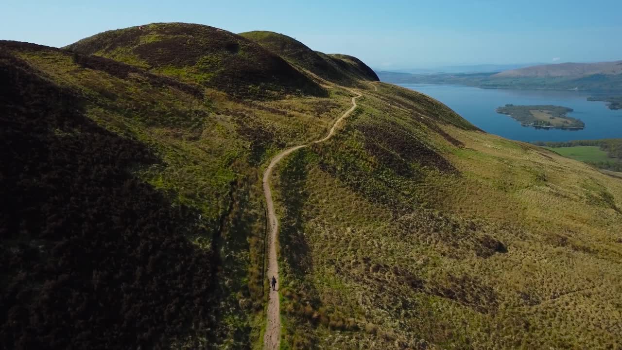 Aerial drone footage gliding over mountainside in Loch Lomond that has a muddy hiking track or road on it during a sunny day while the mountain is covered with green and brown grass and vegetation.