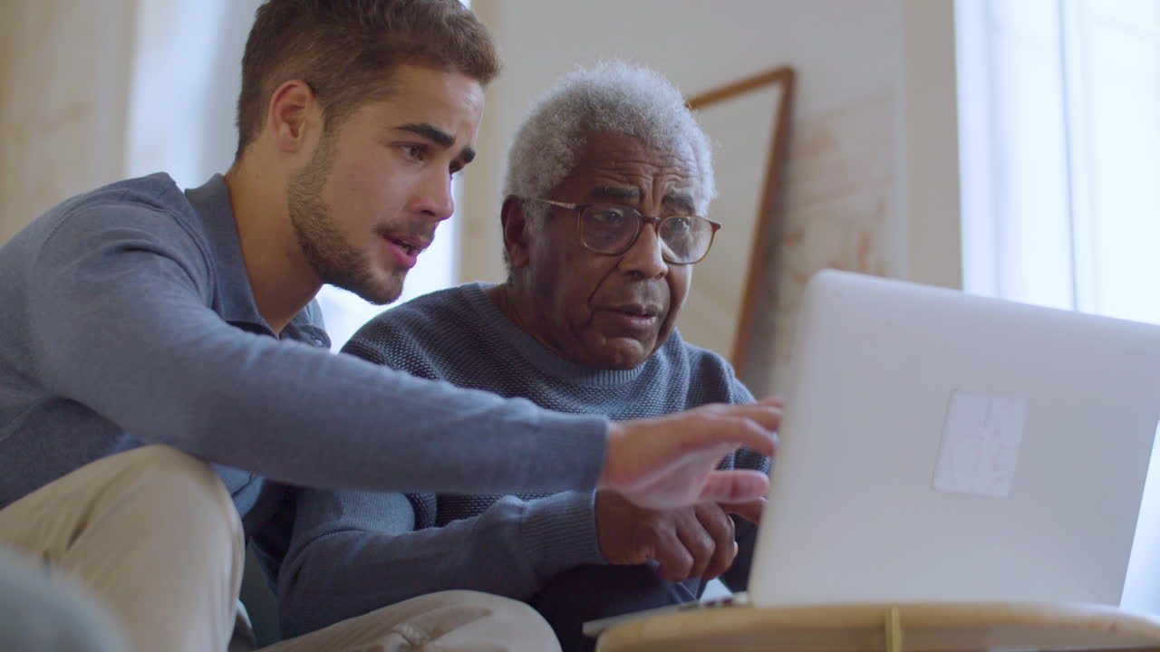 Bearded Caucasian guy helping senior Black man with using laptop