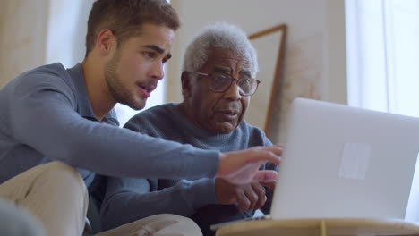 Bärtiger-Kaukasier,-Der-Einem-älteren-Schwarzen-Mann-Bei-Der-Benutzung-Seines-Laptops-Hilft