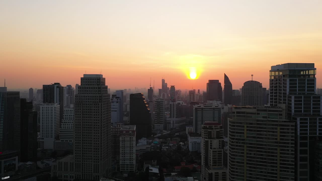 Vista aérea épica de drone voando lateralmente ao lado de edifícios, horizonte e arranha-céus do distrito de Sukhumvit durante um nascer do sol vermelho e colorido em Bangkok, Tailândia.