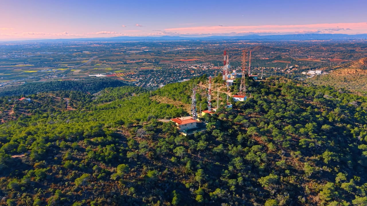Torri di telecomunicazione su una collina che domina una vasta pianura agricola. Veduta aerea di antenne di comunicazione su una cima di montagna con una città e campi sullo sfondo al tramonto