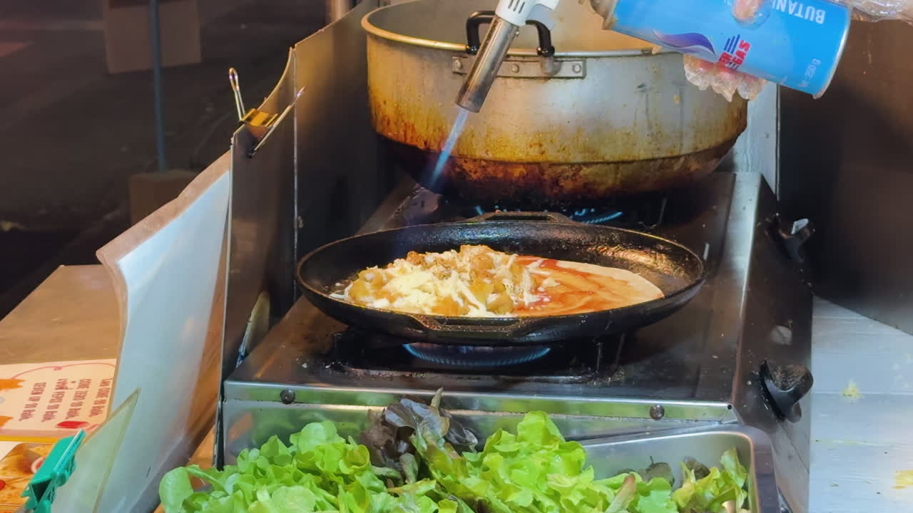 Flame torch browning a dish in Watchediluang Varaviharn kitchen, Chiang Mai, with stainless steel workspace and fresh salad - Thailand