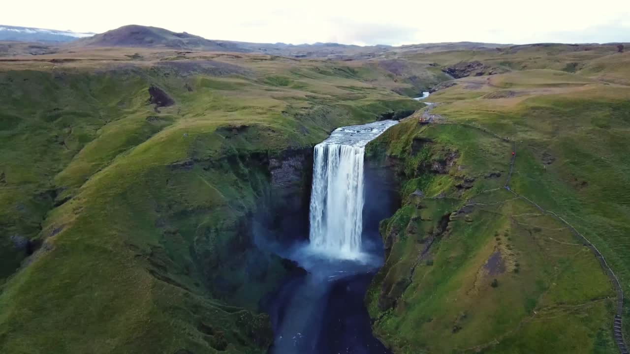 Breathtaking aerial view capturing Skógafoss waterfall cascading down a cliff, creating misty sprays while surrounded by lush green hills and a winding river in Iceland, drone establishing shot