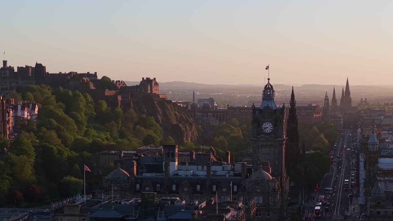 Órbita cinematográfica de drone sobre Edimburgo ao pôr do sol, mostrando o castelo histórico, o relógio da cidade e edifícios majestosos banhados em luz dourada.
