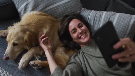 smiling caucasian woman taking selfie with smartphone, stroking her pet dog on sofa next to her