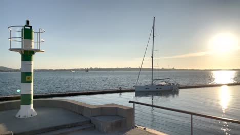 Barco-En-El-Puerto-Deportivo-Con-Un-Faro-Cerca-De-La-Señalización,-El-Reflejo-Del-Sol-En-El-Agua-Del-Mar