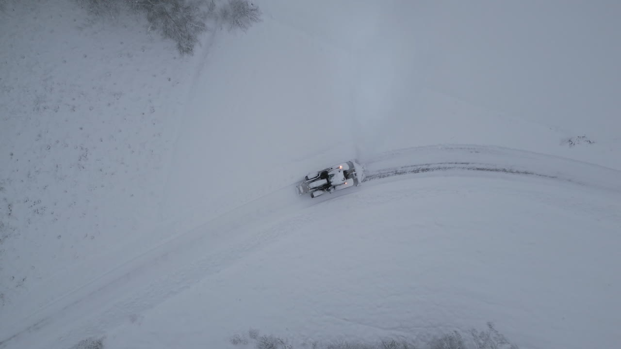 Tractor plowing snow on rural countryside road in winter, ascending top down aerial view