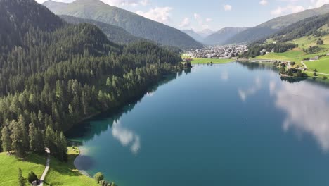 aerial drone flies over serene blue lake water, with green tree filled mountain slopes and town nestled between mountains in davos, kanton graubünden, switzerland