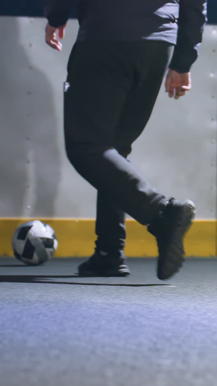 Leg view of man kicking soccer ball against wall under bright stadium lighting at night, focus on athletic motion and shadow with urban environment and illuminated background