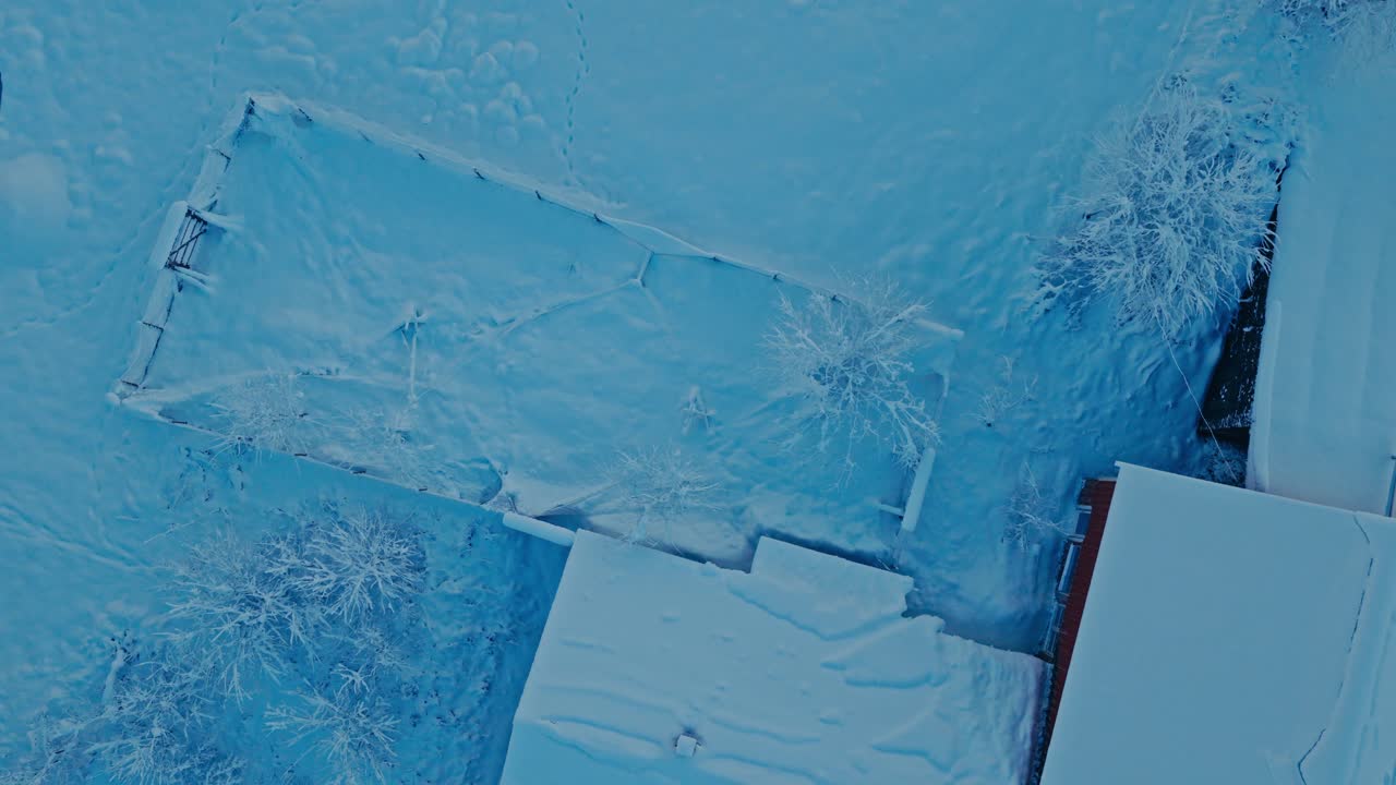 Neve cobre um quintal cercado e telhados, com árvores cobertas de geada delimitando as bordas em uma cena de inverno tranquila - Tomada aérea ascendente