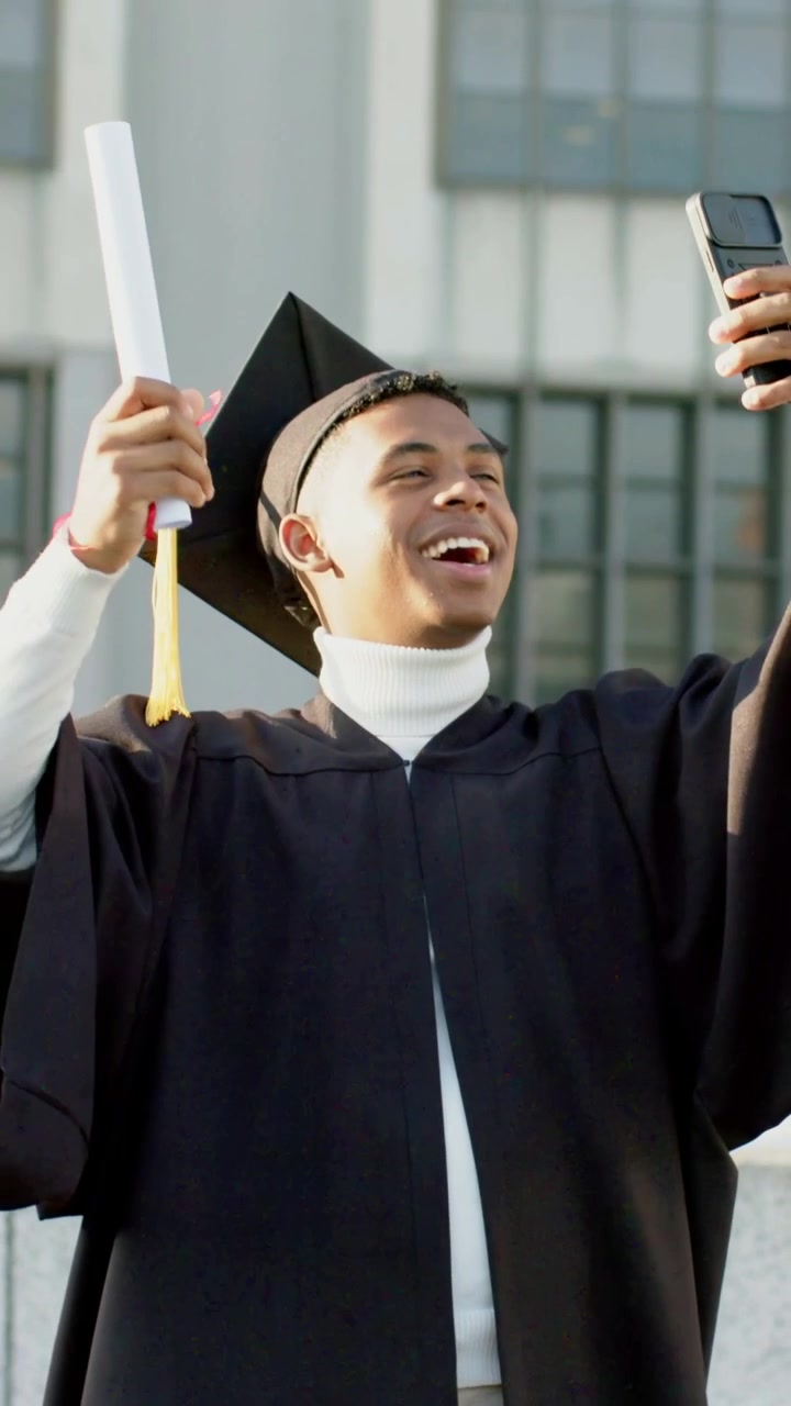 Video verticale: Con il telefono in mano, un laureato in toga e tocco filma una celebrazione nel campus, sventolando il diploma.