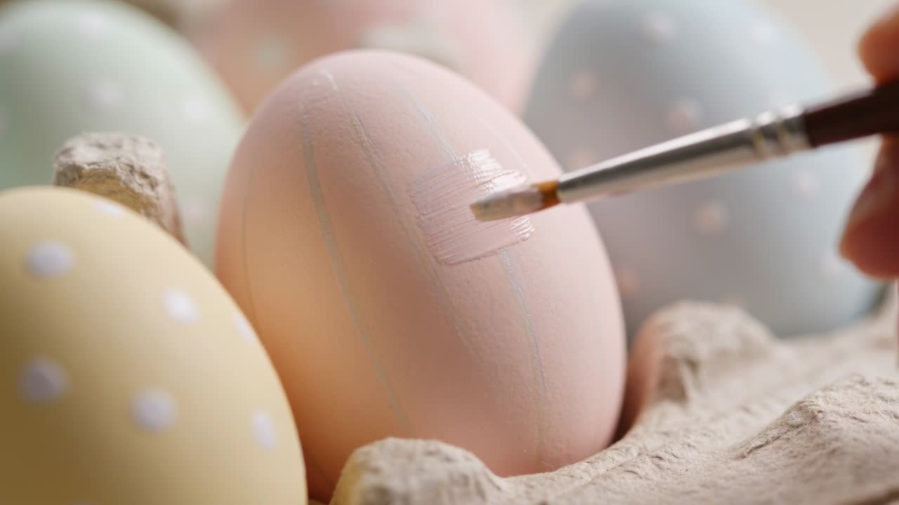 Hand holding brush entering from right, painting pastel pink egg with white strokes for decoration