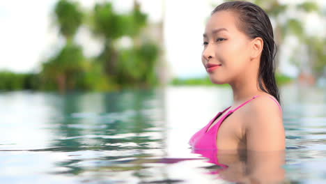 dreamy side view scene of a beautiful woman standing in the swimming pool during a bright sunny day, looking around and smiling with joy on her face