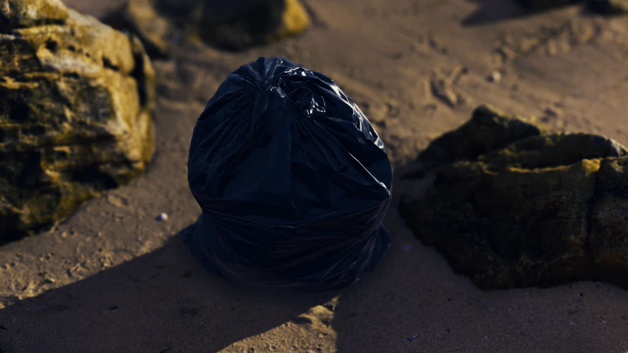 Trash bag left on sandy beach near rocks under morning sunlight