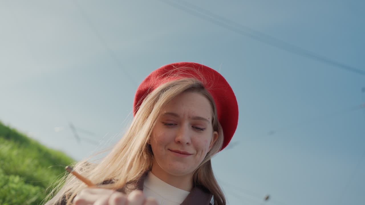 Ung kvinde i rød baret skriver i notesbog på græsklædt skråning under klar blå himmel med telefonpæle, blødt sollys, filmisk nærbillede, der viser fokuserede blyantstrøg og eftertænksom stemning